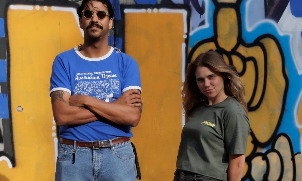 Vor einer gelb-blauen Graffitiwand stehen Natalie und Greg von Press Club. Natalie hält lässig zurückgelehnt die Hände hinterm Rücken und Greg trägt eine Sonnenbrille.