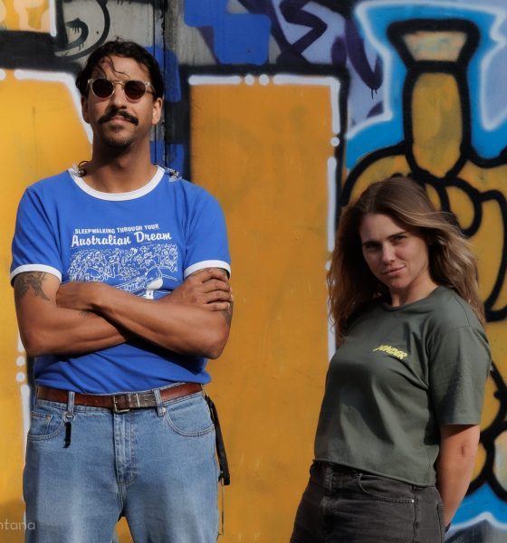 Vor einer gelb-blauen Graffitiwand stehen Natalie und Greg von Press Club. Natalie hält lässig zurückgelehnt die Hände hinterm Rücken und Greg trägt eine Sonnenbrille.