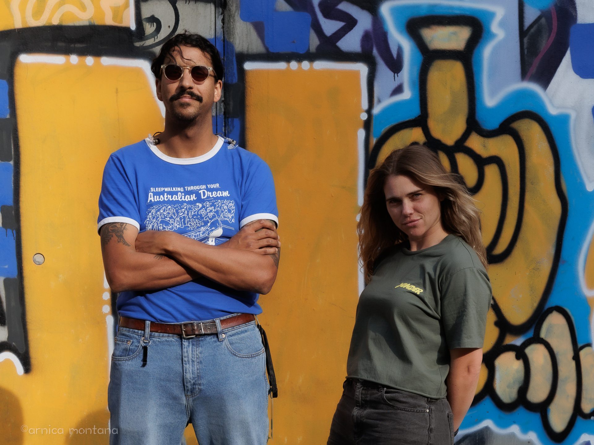 Vor einer gelb-blauen Graffitiwand stehen Natalie und Greg von Press Club. Natalie hält lässig zurückgelehnt die Hände hinterm Rücken und Greg trägt eine Sonnenbrille.