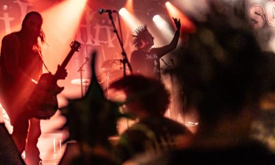 Eine lebendige Konzertszene mit rötlichen Lichtern. auf der Bühne sind mehrere Musiker zu erkennen, im Vordergrund die Personen sind unscharf und haben Spike-Frisuren.