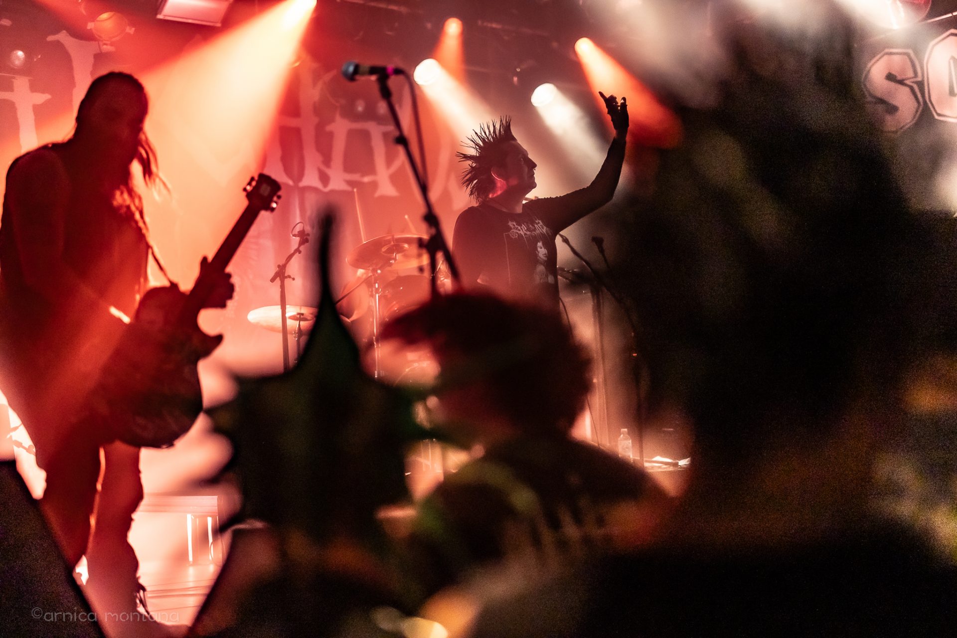 Eine lebendige Konzertszene mit rötlichen Lichtern. auf der Bühne sind mehrere Musiker zu erkennen, im Vordergrund die Personen sind unscharf und haben Spike-Frisuren.