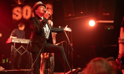 Eine Frau mit Pork Pie Hut in schwarz mit weißem Hutband, schwarzem Jacket und schwarzen Cigarette Pants steht mit einem Schuh auf dem Monitor und singt zum Publikum. Eine Hand macht eine einladenden Bewegung, die andere Hand hält das. Mikrofon, während sie singt.