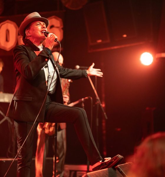 Eine Frau mit Pork Pie Hut in schwarz mit weißem Hutband, schwarzem Jacket und schwarzen Cigarette Pants steht mit einem Schuh auf dem Monitor und singt zum Publikum. Eine Hand macht eine einladenden Bewegung, die andere Hand hält das. Mikrofon, während sie singt.