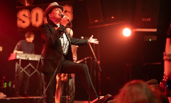 Eine Frau mit Pork Pie Hut in schwarz mit weißem Hutband, schwarzem Jacket und schwarzen Cigarette Pants steht mit einem Schuh auf dem Monitor und singt zum Publikum. Eine Hand macht eine einladenden Bewegung, die andere Hand hält das. Mikrofon, während sie singt.