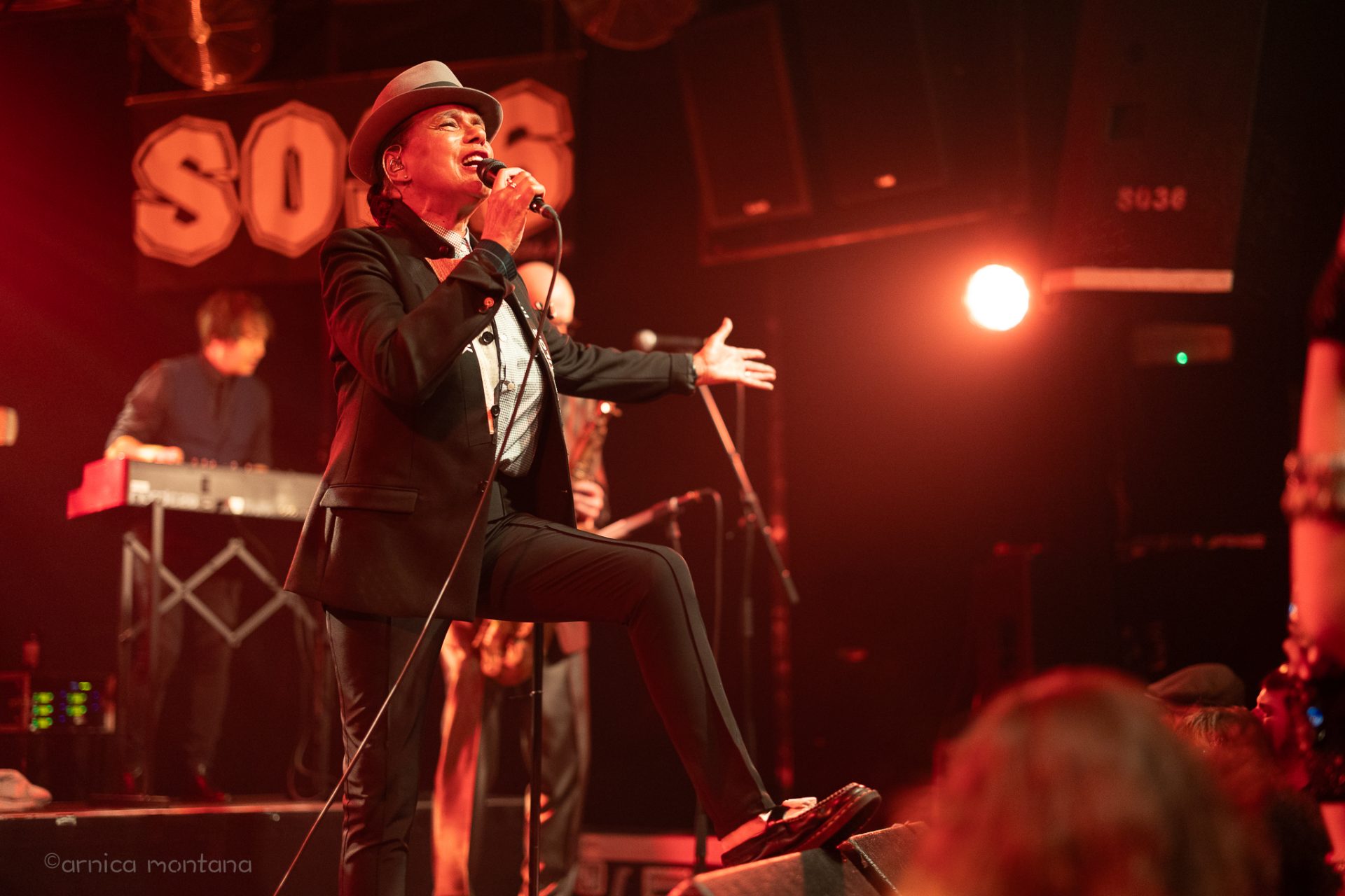 Eine Frau mit Pork Pie Hut in schwarz mit weißem Hutband, schwarzem Jacket und schwarzen Cigarette Pants steht mit einem Schuh auf dem Monitor und singt zum Publikum. Eine Hand macht eine einladenden Bewegung, die andere Hand hält das. Mikrofon, während sie singt.